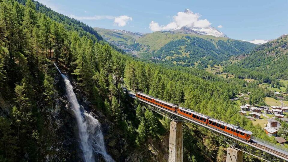 A train of Gornergrat railway travels on a bridge over a deep gorge and Findelbach Waterfall tumbles down the cliff with majestic Matterhorn Mountain in background, in Zermatt, Valais, Switzerland